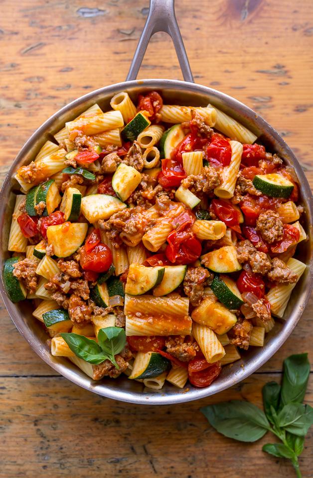 Skillet of rigatoni with tomato sauce, sausage, and zucchini on brown table.
