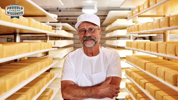 A man in a white apron and hat stands in a room full of shelves stacked with cheese.
